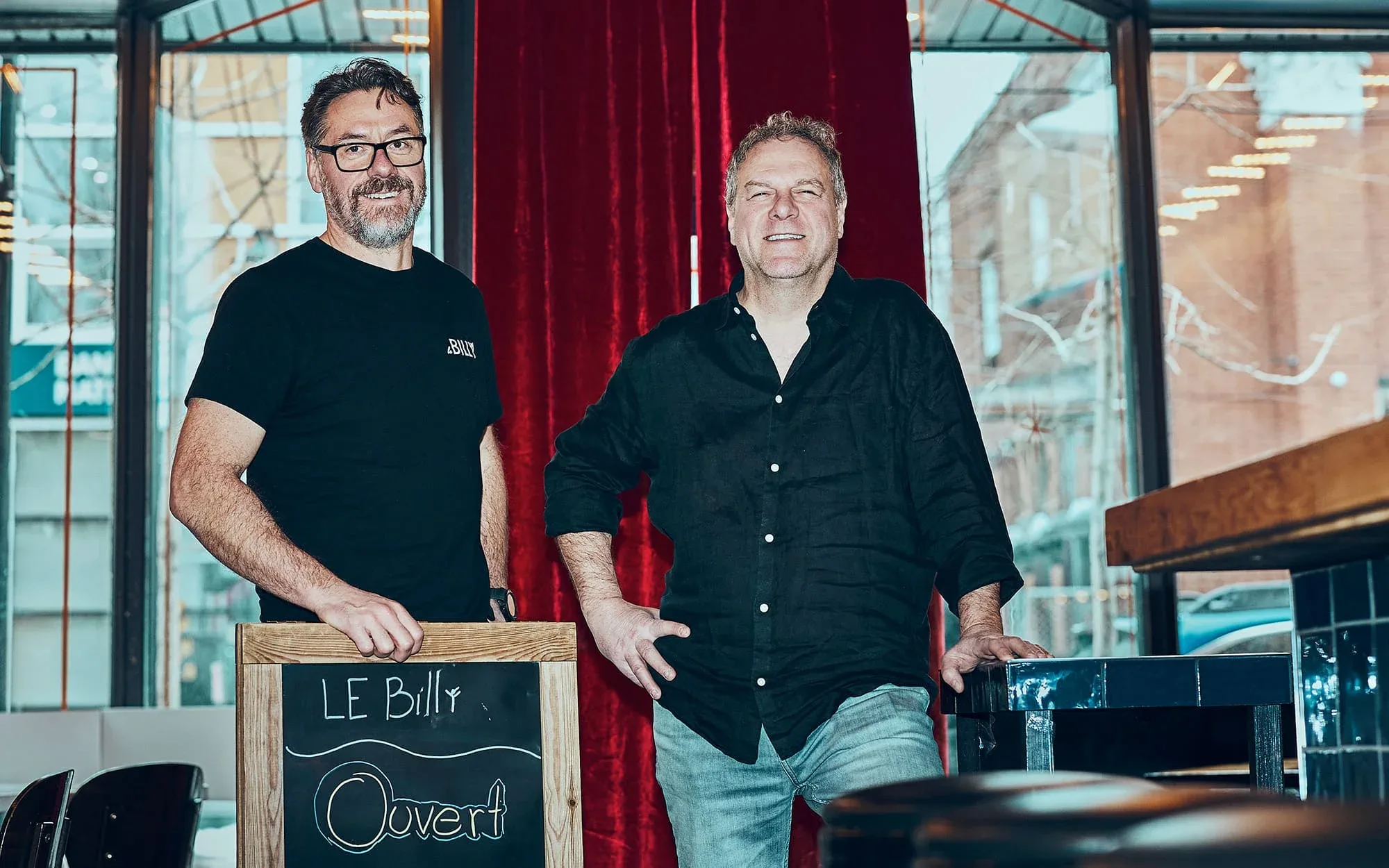Two men in the middle of a bar looking at the camera, with a sign in front of them stating that Billy's is open