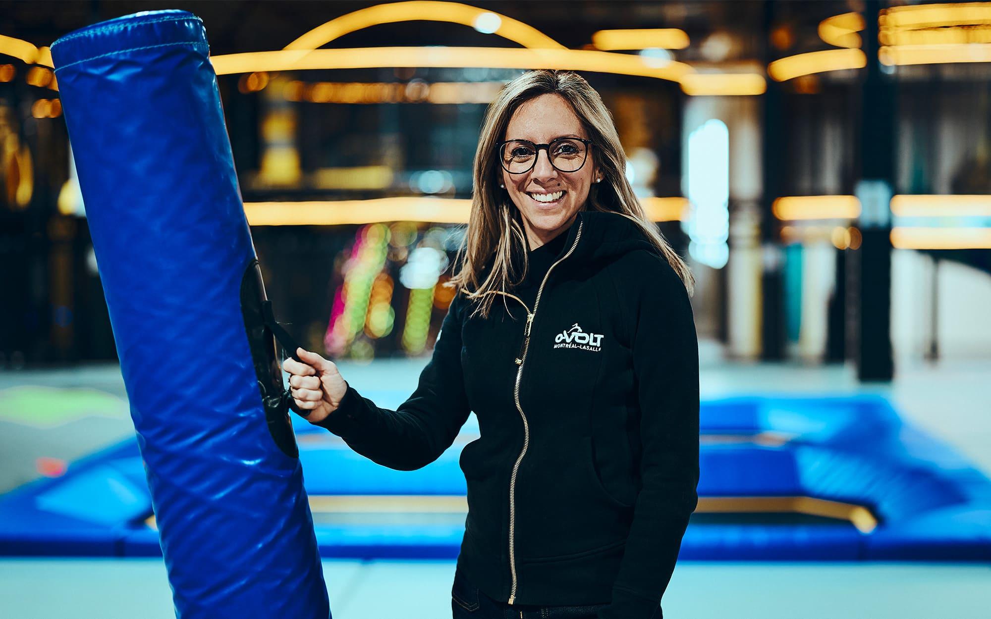 Femme à l'intérieur d'un bâtiment devant des trampolines qui sourit devant la caméra en tenant un coussin protecteur